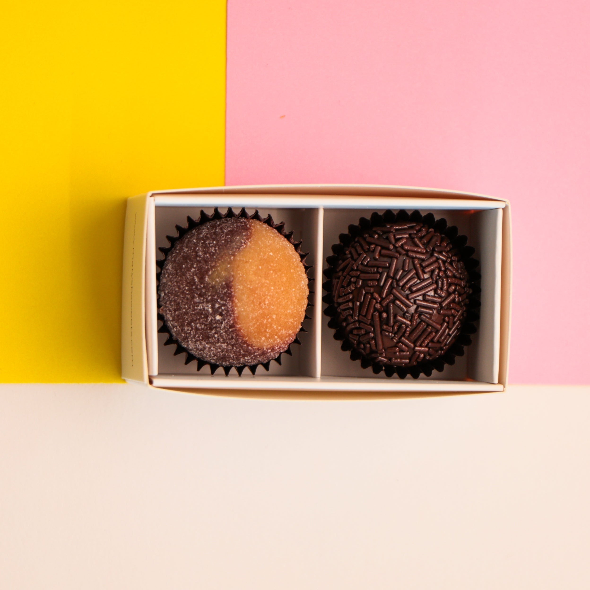 Two chocolates brigadeiros in a gift box on a pink, yellow background.