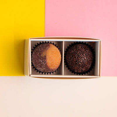Two chocolates brigadeiros in a gift box on a pink, yellow background.