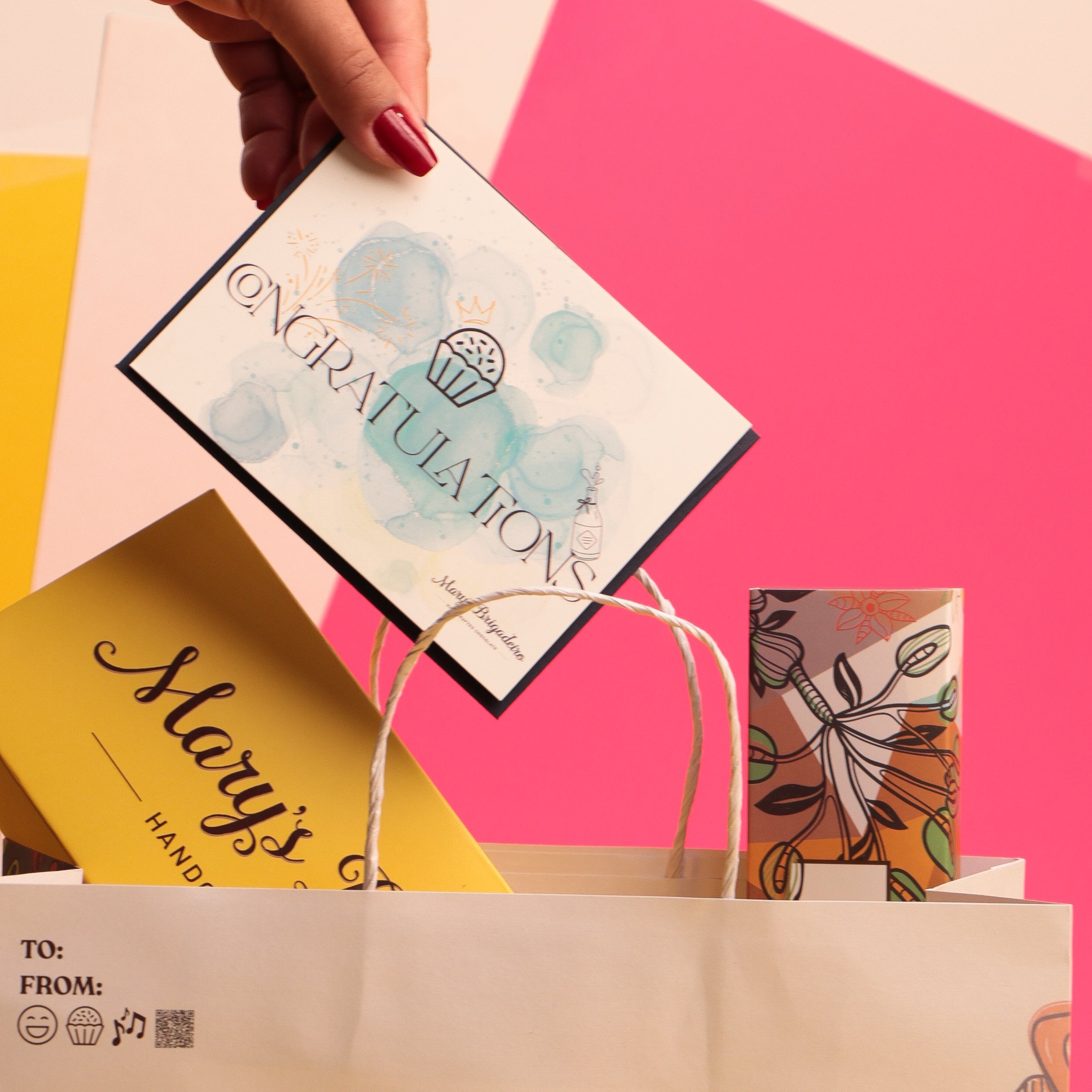 Hand holding a 'Congratulations' card with a colorful background featuring other cards and a branded chocolate bag.